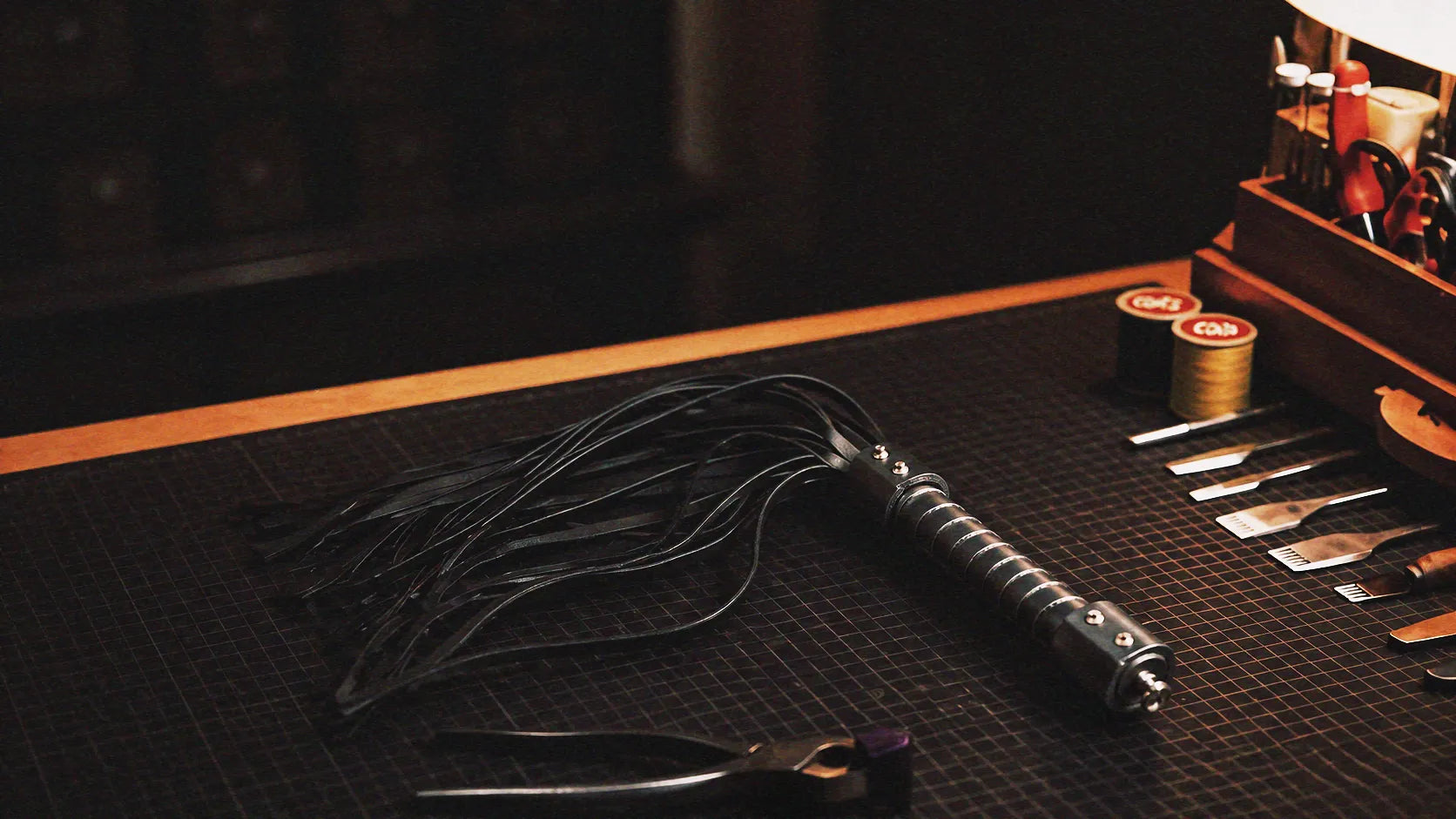 Midnight Discipline Leather Flogger warm tabletop studio scene with wooden box, matte black leather, and slim tails.