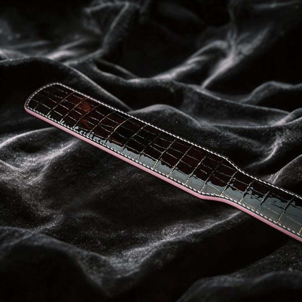 Noir Grid Patent Texture Leather Paddle angled on black satin, showing black patent leather and grid texture.