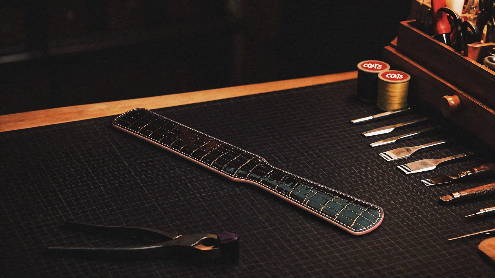 Noir Grid Patent Texture Leather Paddle warm tabletop studio scene with wooden box, black patent leather, and grid texture.
