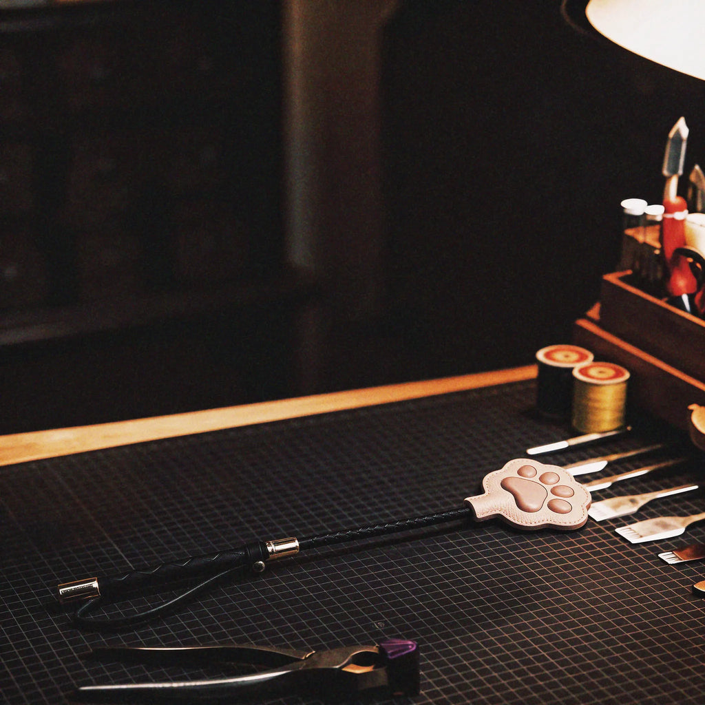 Paw Whisper cat paw paddle workbench scene with the blush paw head, braided handle, and slim black shaft on a gridded studio mat