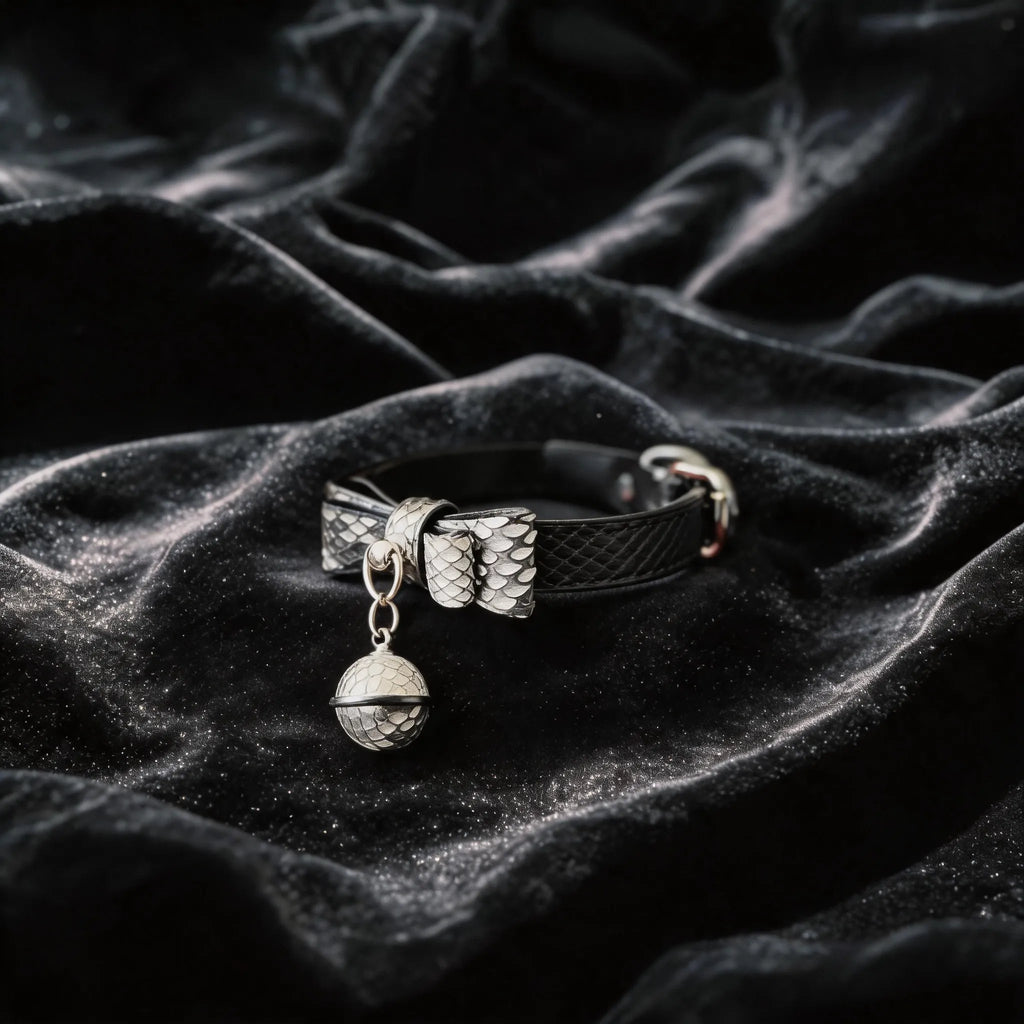 Serpentine Grace Bow Bell Leather Collar angled on black satin, showing black and white python-textured leather and bow and bell charm.
