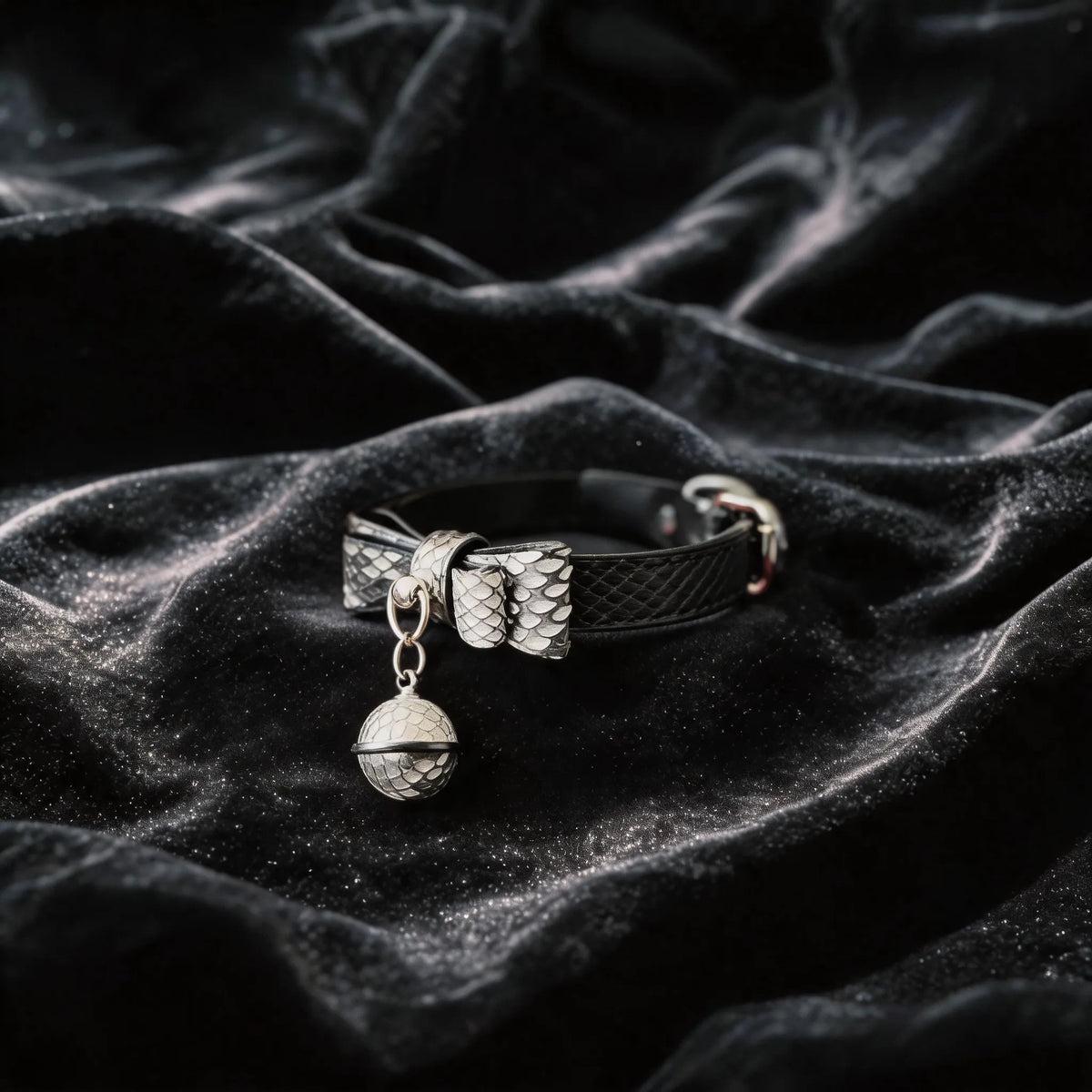 Serpentine Grace Bow Bell Leather Collar angled on black satin, showing black and white python-textured leather and bow and bell charm.