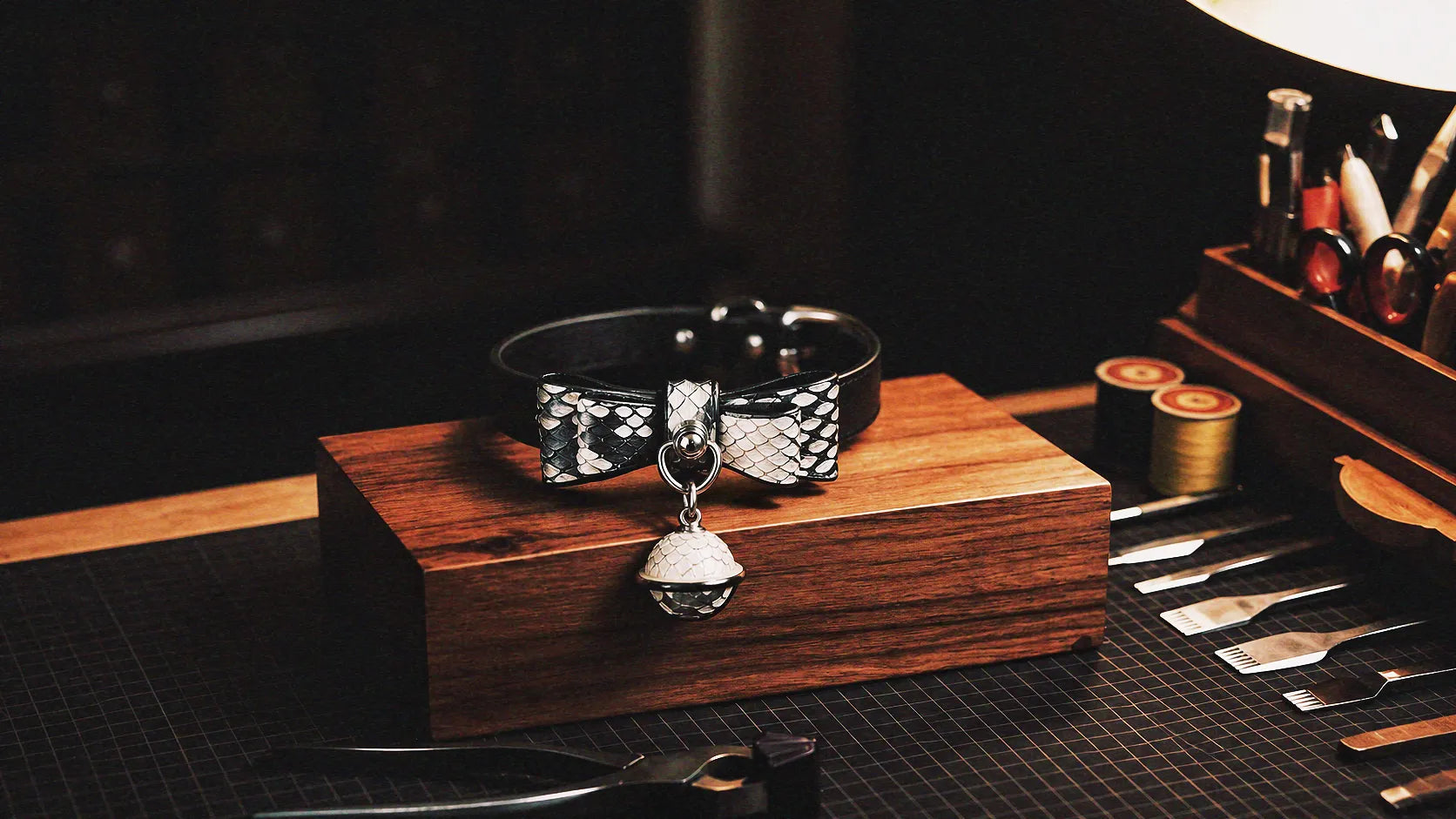 Serpentine Grace Bow Bell Leather Collar warm tabletop studio scene with wooden box, black and white python-textured leather, and bow and bell charm.